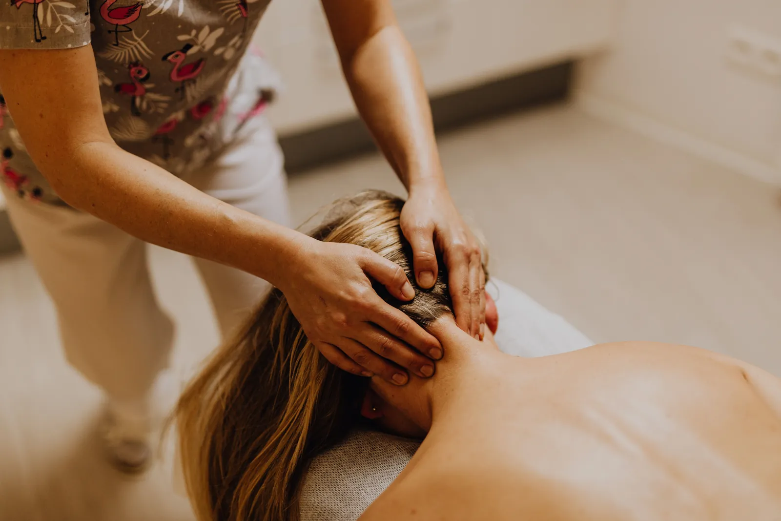 A massage therapist working the back of a client's neck with both hands, the client lying face down on a treatment table